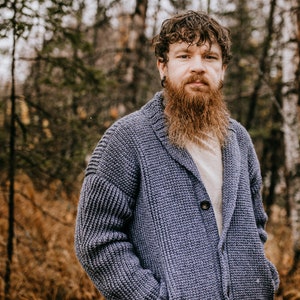 May include: A man with a long, red beard is wearing a blue knitted cardigan with a shawl collar and a single button closure. He is standing in a forest setting with trees and brown foliage in the background.