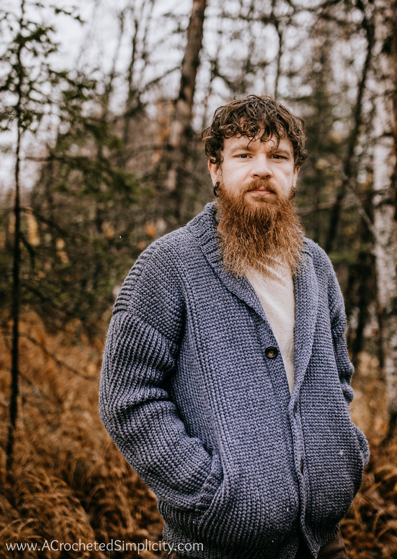 May include: A man with a long, red beard is wearing a blue knitted cardigan with a shawl collar and a single button closure. He is standing in a forest setting with trees and brown foliage in the background.