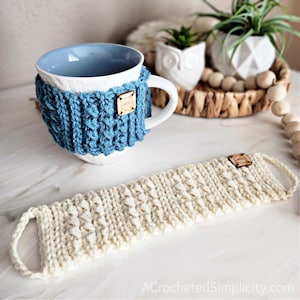 May include: A white ceramic mug with a blue crocheted cosy. The cosy has a wooden tag with the word "Cozy" on it. A white crocheted mug cosy with a wooden tag is also shown.