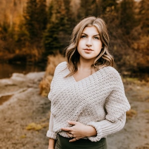 May include: A woman wearing a light beige crocheted sweater with a V-neck and a dark green skirt. She is standing in a forest setting with trees in the background.
