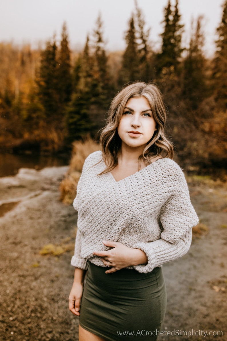 May include: A woman wearing a light beige crocheted sweater with a V-neck and a dark green skirt. She is standing in a forest setting with trees in the background.