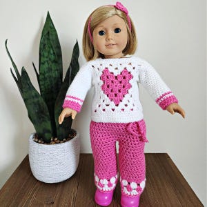 Peut inclure: Une poupée portant un pull en crochet blanc avec un motif de cœur rose et un pantalon en crochet rose avec des accents blancs. La poupée a les cheveux blonds et les yeux bleus.