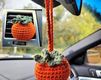 Puede incluir: Una calabaza naranja de ganchillo con hojas verdes colgando del espejo retrovisor de un coche. La calabaza también se muestra en primer plano colgando de la ventilación de un coche.