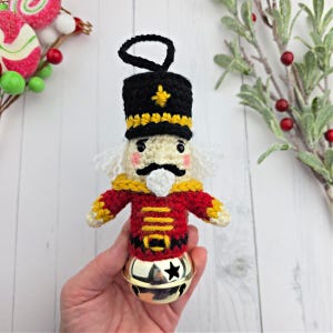 May include: A handmade crochet Christmas ornament in the shape of a nutcracker soldier. The ornament features a black hat with a gold star, a red jacket with gold accents, and a gold bell base. The ornament is approximately 4 inches tall.