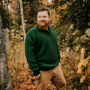 Pu&ograve; includere: Un maglione verde lavorato a maglia con colletto a scialle e polsini e orlo a coste &egrave; indossato con pantaloni color cachi. L'immagine &egrave; ambientata in una foresta con fogliame autunnale. Il testo "ACrochetedSimplicity" &egrave; in basso.