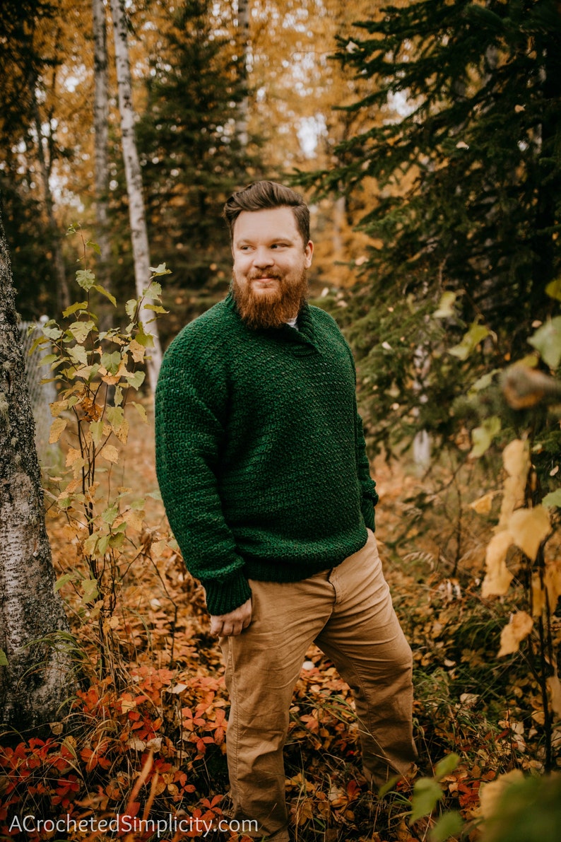Pu&ograve; includere: Un maglione verde lavorato a maglia con colletto a scialle e polsini e orlo a coste &egrave; indossato con pantaloni color cachi. L'immagine &egrave; ambientata in una foresta con fogliame autunnale. Il testo "ACrochetedSimplicity" &egrave; in basso.
