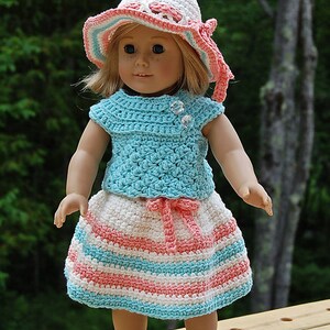 May include: A doll wearing a crocheted outfit in shades of pink, white, and blue. The outfit includes a sleeveless top, a skirt with stripes, and a matching hat.