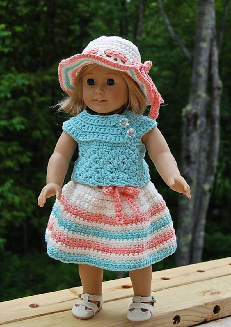 May include: A doll wearing a crocheted outfit in shades of pink, white, and blue. The outfit includes a sleeveless top, a skirt with stripes, and a matching hat.