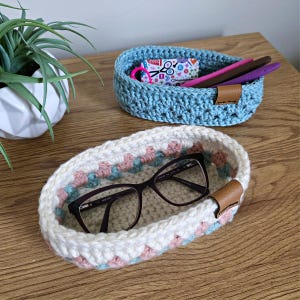 May include: Two handmade crochet baskets in shades of blue and white with pink and teal accents. One basket holds eyeglasses, the other holds pens and a small patterned box. Both have brown leather tags.