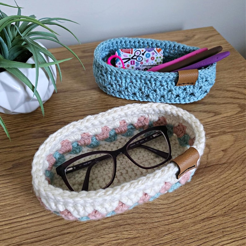 May include: Two crocheted oval baskets with leather handles. One basket is light blue and the other is white with pink and green accents. The white basket has a pair of eyeglasses inside.