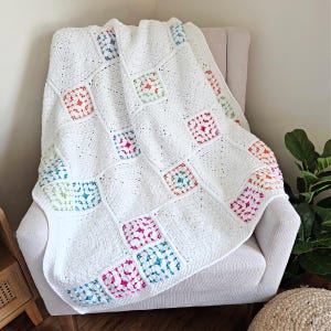 Puede incluir: Una manta blanca de ganchillo sobre una silla. La manta presenta un patrón de cuadrados coloridos en tonos de azul, verde, naranja y rosa. La manta está hecha de un hilo suave y texturizado y parece hecha a mano.