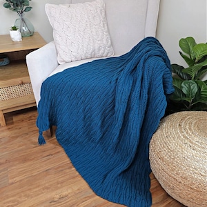 May include: A teal blue knitted throw blanket with a textured pattern and tassels draped over a white armchair. A white cable-knit pillow rests on the chair. The scene includes a wooden side table and a woven ottoman.