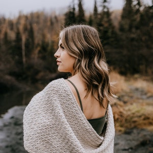 May include: A woman wearing a light gray crochet shawl with a green tank top underneath. The shawl is draped over her shoulders and is open in the back. She is standing in front of a blurred background of trees.