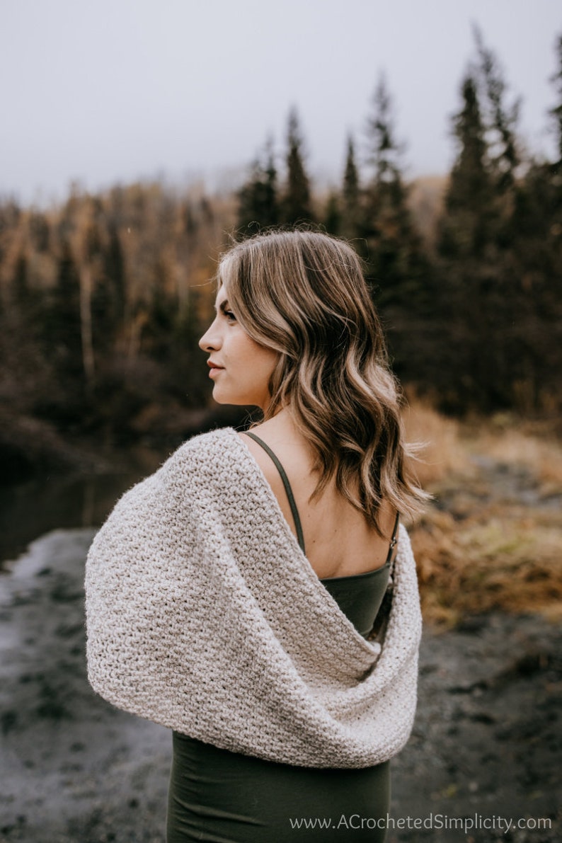 May include: A woman wearing a light gray crochet shawl with a green tank top underneath. The shawl is draped over her shoulders and is open in the back. She is standing in front of a blurred background of trees.