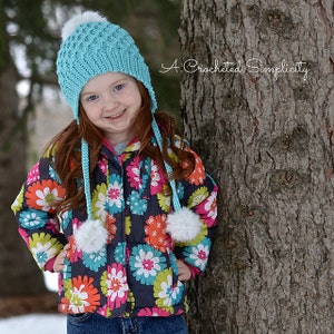 May include: A young girl wearing a blue crocheted hat with white pom poms and matching ear flaps. She is wearing a colorful floral print jacket and blue jeans. The image is captioned "A Crocheted Simplicity".