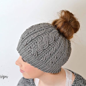 May include: Gray crochet beanie with a braided design, worn with a messy bun hairstyle.