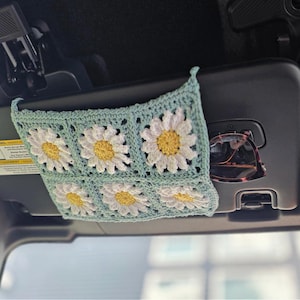 May include: A crocheted car visor organizer in a light blue hue, adorned with daisy motifs in white and yellow. The organizer is designed to hold items and is attached to the car's visor. Sunglasses are visible in the visor's storage compartment.