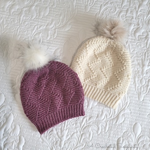 Puede incluir: Dos gorros de ganchillo con pompones. Un gorro es de color morado oscuro y el otro es de color crema.