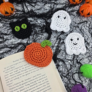 Puede incluir: Decoraciones de Halloween hechas a mano con ganchillo. La imagen muestra un gato negro con ojos verdes, dos fantasmas blancos y una calabaza naranja con un tallo verde, dispuestos sobre una tela de malla negra.