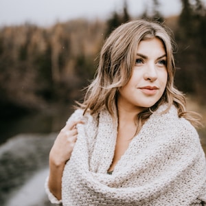 May include: A woman wearing a light beige crochet scarf with a loose, draping style. The scarf is worn over a dark green top. The woman is looking off to the side with a thoughtful expression.