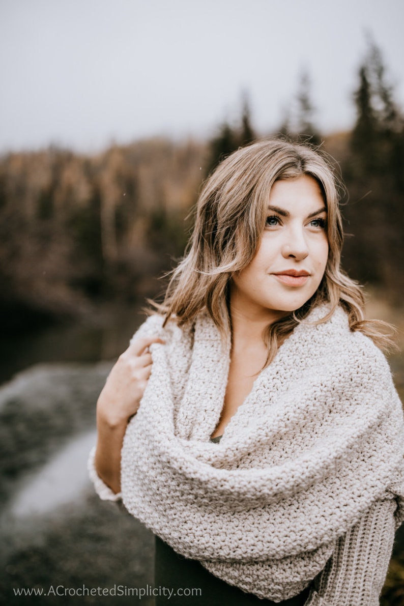 May include: A woman wearing a light beige crochet scarf with a loose, draping style. The scarf is worn over a dark green top. The woman is looking off to the side with a thoughtful expression.