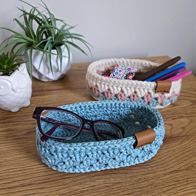 May include: Two crocheted oval baskets, one blue and one with pink, white, and green stripes. The blue basket has a pair of glasses inside and a brown leather handle. The striped basket has a brown leather handle.