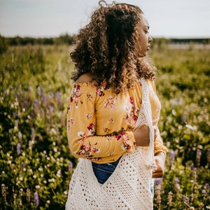 May include: A woman wearing a yellow floral off-the-shoulder top and denim shorts is holding a white crocheted tote bag. The bag has a large, open top and a long, fringed strap. She is standing in a field of wildflowers.