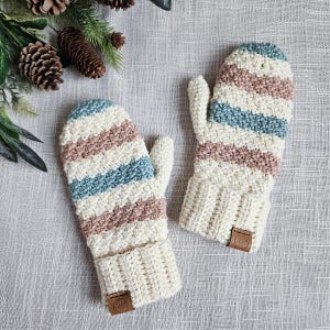 May include: A pair of crocheted mittens with a striped pattern in shades of beige, blue, and white. The mittens have a textured, fluffy look and are made with a soft yarn.
