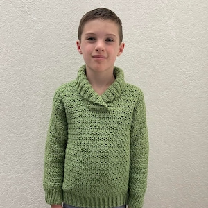 May include: A young person wearing a green knitted sweater with a collared neckline. The sweater has a textured stitch pattern.