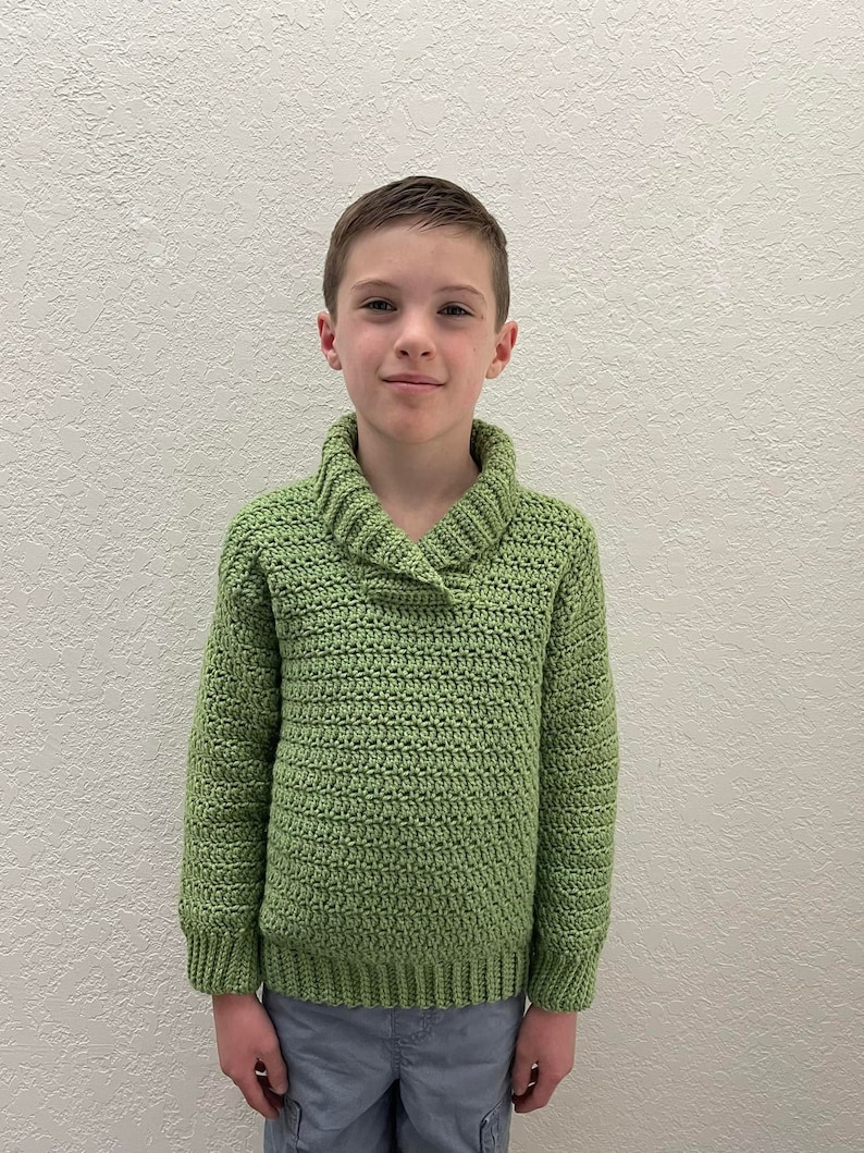 May include: A young person wearing a green knitted sweater with a collared neckline. The sweater has a textured stitch pattern.