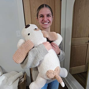 Puede incluir: Un gran peluche de perro de ganchillo hecho a mano en blanco, beige y marr&oacute;n. El perro tiene orejas ca&iacute;das, una mancha marr&oacute;n en la cabeza y una cola. Una persona sostiene el peluche.