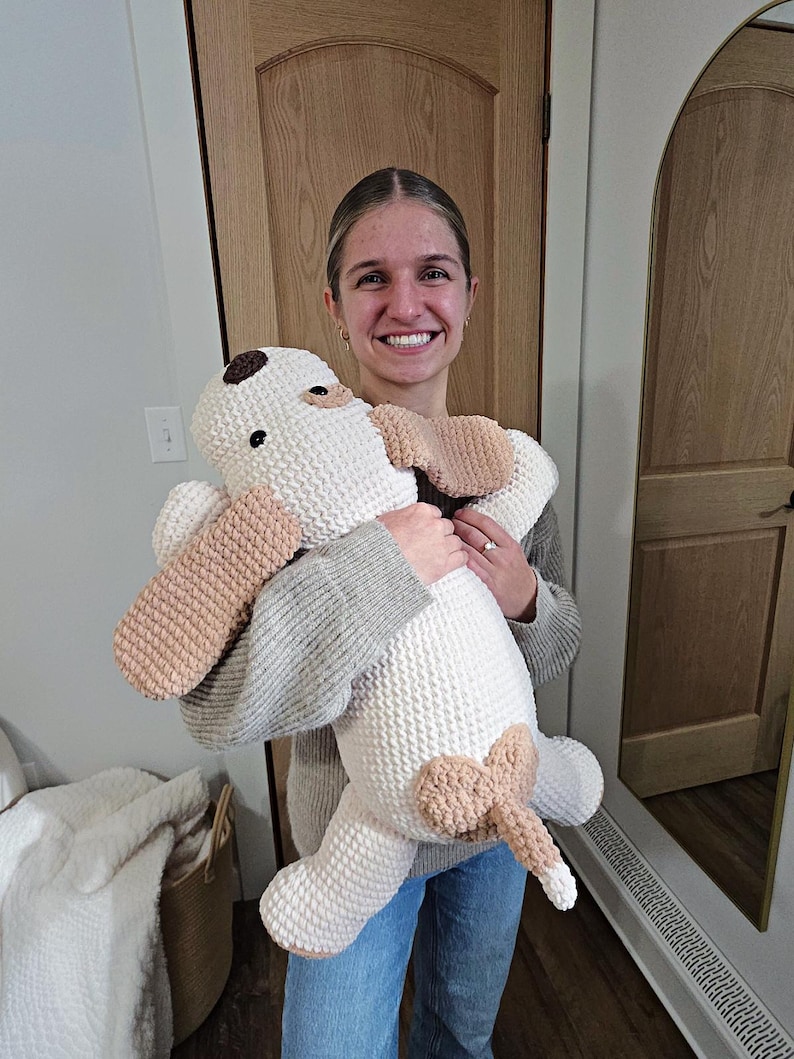 Puede incluir: Un gran peluche de perro de ganchillo hecho a mano en blanco, beige y marr&oacute;n. El perro tiene orejas ca&iacute;das, una mancha marr&oacute;n en la cabeza y una cola. Una persona sostiene el peluche.