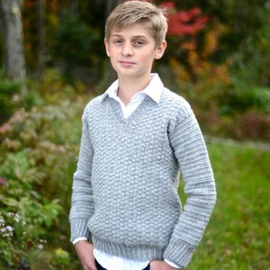 May include: A gray crocheted sweater with a V-neck and long sleeves. The sweater is worn by a person standing in front of a blurred background of trees.