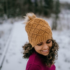 May include: A woman wearing a brown crocheted beanie with a faux fur pom pom and a burgundy sweater. The beanie has a textured stitch pattern. The woman is standing in a snowy outdoor setting.