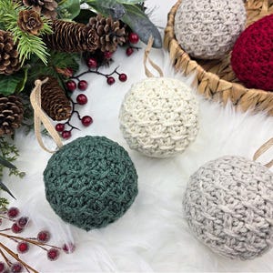 May include: Handmade crochet Christmas baubles in green, cream, grey, and red. Each ball-shaped decoration has a textured pattern and a tan ribbon for hanging. Some baubles are displayed in a woven basket, with festive foliage.