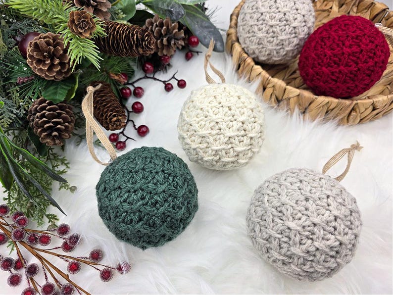 May include: Handmade crochet Christmas baubles in green, cream, grey, and red. Each ball-shaped decoration has a textured pattern and a tan ribbon for hanging. Some baubles are displayed in a woven basket, with festive foliage.