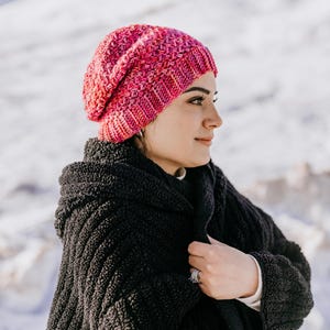 May include: A pink and red knitted beanie hat. The hat is made of a textured yarn and has a ribbed brim. The person is wearing a black coat.