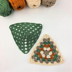 May include: Two handmade crochet granny squares in a triangle shape. One is solid green, the other is a mix of beige, brown, green, and orange. Several balls of yarn in shades of green, orange, beige, and brown are in the background.