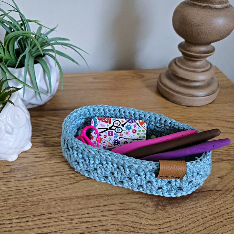 May include: A light blue crocheted oval-shaped basket with a brown leather handle. The basket is filled with craft supplies, including a pair of pink scissors, a pink and white card with sewing-themed designs, and three purple, pink, and brown crochet hooks.