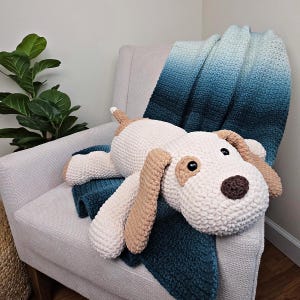 Könnte beinhalten: Ein großer, handgefertigter Häkelhund-Plüschtier in Creme, Beige und Braun, der auf einer teal- und blau-Ombre-Decke liegt. Der Hund hat Schlappohren, eine braune Nase und schwarze Augen. Die Decke ist über einen hellgrauen Sessel drapiert.