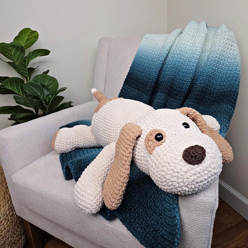 Puede incluir: Un gran peluche de perro de ganchillo hecho a mano en crema, marr&oacute;n y marr&oacute;n, descansando sobre una manta degradada en azul y azul. El perro tiene orejas ca&iacute;das, una nariz marr&oacute;n y ojos negros. La manta est&aacute; sobre un sill&oacute;n gris claro.