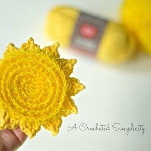 May include: A crocheted yellow sun with a textured center, held in a hand. The sun is made with yarn and has a crocheted design.
