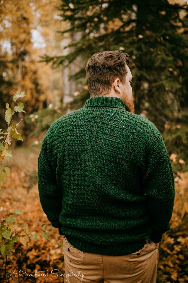 Pu&ograve; includere: Un maglione all'uncinetto verde scuro con collo alto e motivo a punto strutturato. La persona indossa il maglione all'aperto in un ambiente forestale con foglie autunnali.