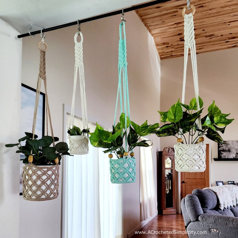 May include: Four macrame plant hangers with different colored pots and plants hanging from a black metal rod. The hangers are made of white and beige yarn. The pots are white, beige, and blue. The plants are green and leafy.