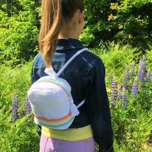 May include: A white crocheted backpack with a rainbow stripe design. The backpack has a small tassel on the bottom. The backpack is being worn by a person in a denim jacket and shorts.