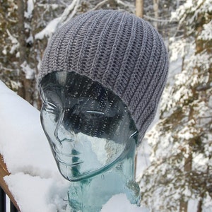 K&ouml;nnte beinhalten: Eine graue H&auml;kelm&uuml;tze auf einem Glas-Mannequin-Kopf. Die M&uuml;tze hat eine gerippte Textur und ist vor einem schneebedeckten Hintergrund platziert.