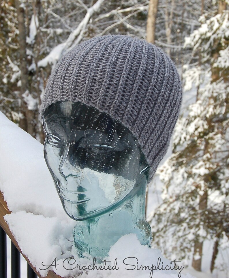 K&ouml;nnte beinhalten: Eine graue H&auml;kelm&uuml;tze auf einem Glas-Mannequin-Kopf. Die M&uuml;tze hat eine gerippte Textur und ist vor einem schneebedeckten Hintergrund platziert.