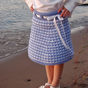 Peut inclure: Une jeune fille portant une chemise blanche et une jupe en crochet gingham bleu et blanc avec un ruban blanc noué autour de la taille. Elle porte des sandales blanches avec des fleurs.