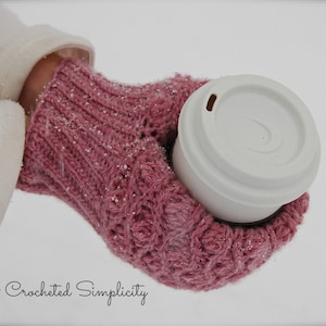 May include: A pair of pink crocheted mittens with a white cup with a lid in the palm of the mittens. The mittens and cup are covered in snow.