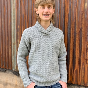 Pu&ograve; includere: Un maglione all'uncinetto grigio chiaro con collo a scialle. Il maglione ha maniche lunghe e orlo e polsini a coste. La persona indossa jeans blu. Lo sfondo &egrave; un muro di metallo arrugginito.